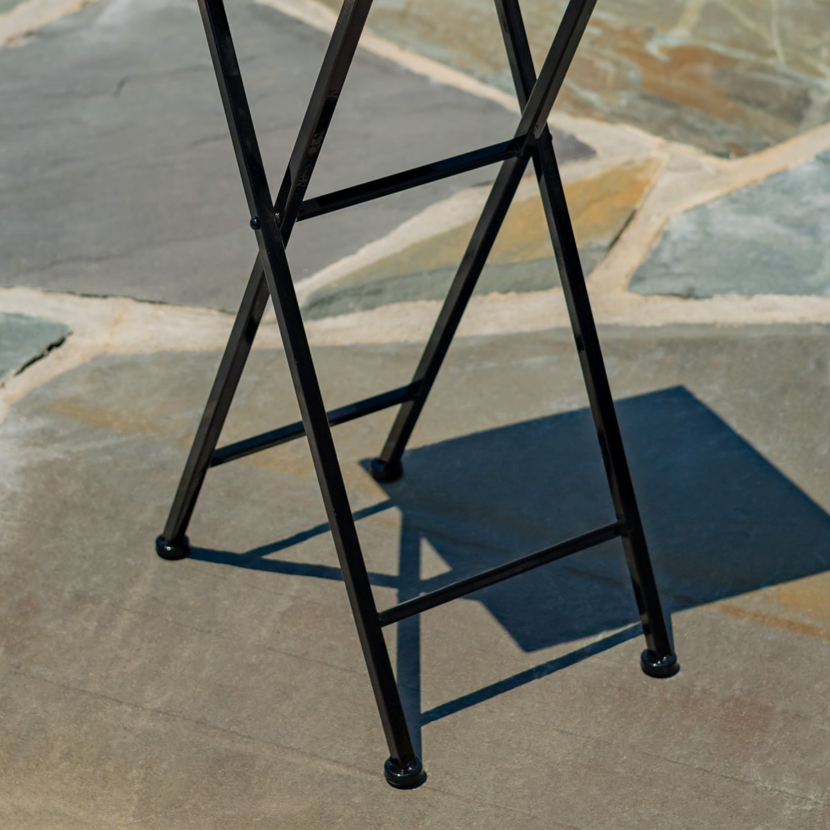 Black metal folding stand legs cross beneath the tabletop on a stone patio.