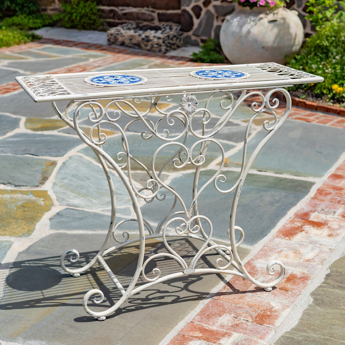 White iron table with a rectangular top featuring two blue mosaic medallions, set on a stone patio.