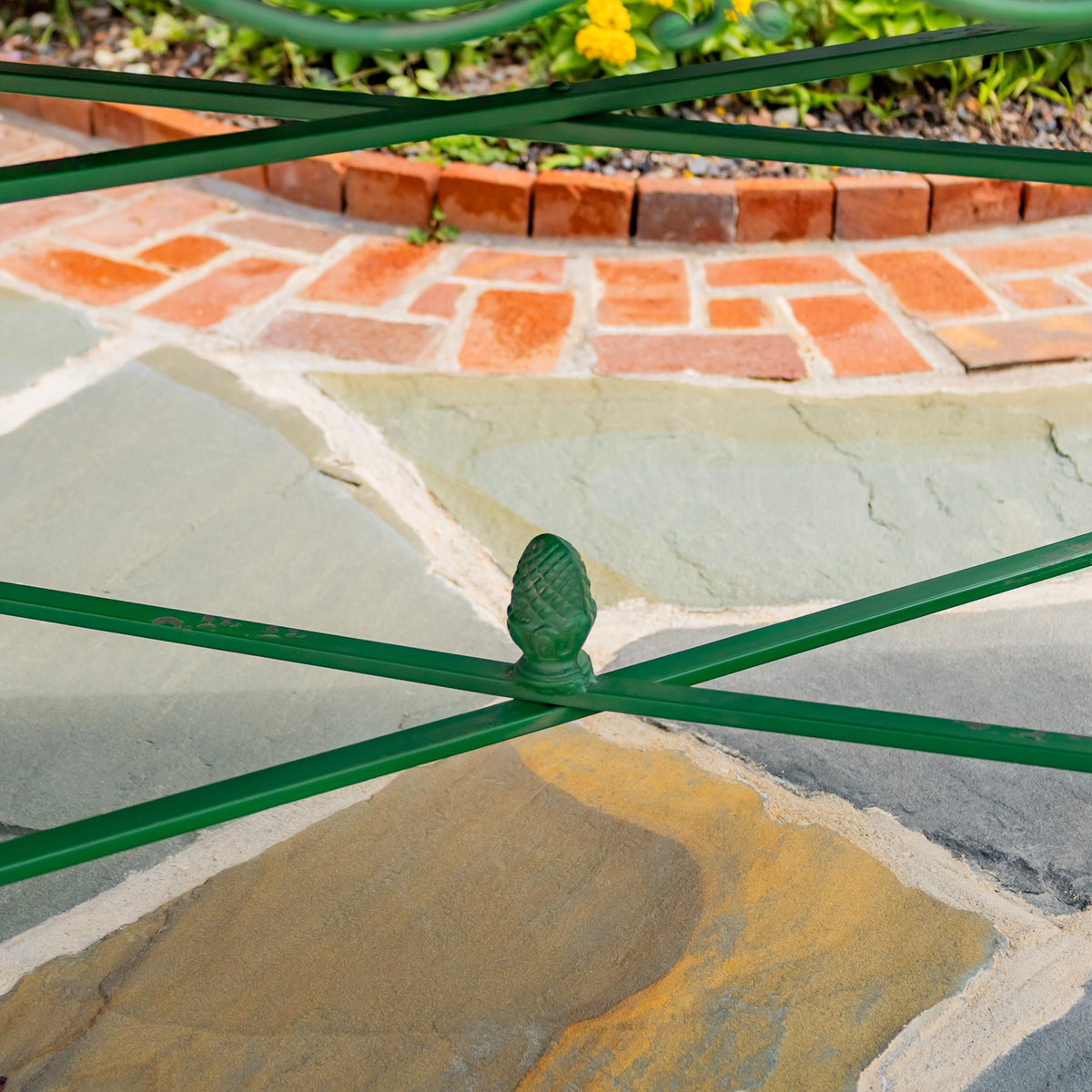 Crossed green metal table supports meet at a decorative finial above a stone patio.
