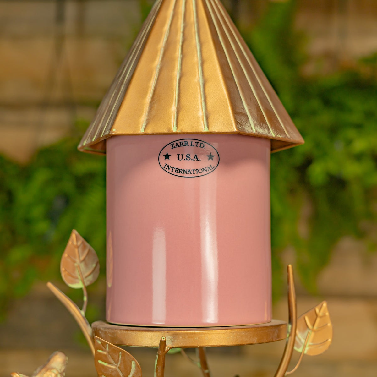 Close-up of the pink birdhouse body, gold-toned roof, and decorative metal leaves and branches.