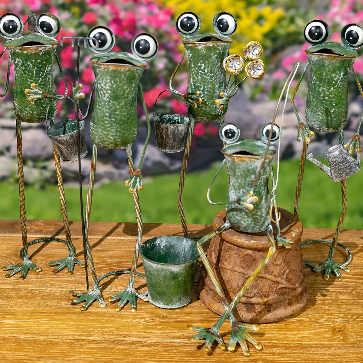 A group of metal frog figures with long legs and small pots, arranged on a wooden surface among flowers.