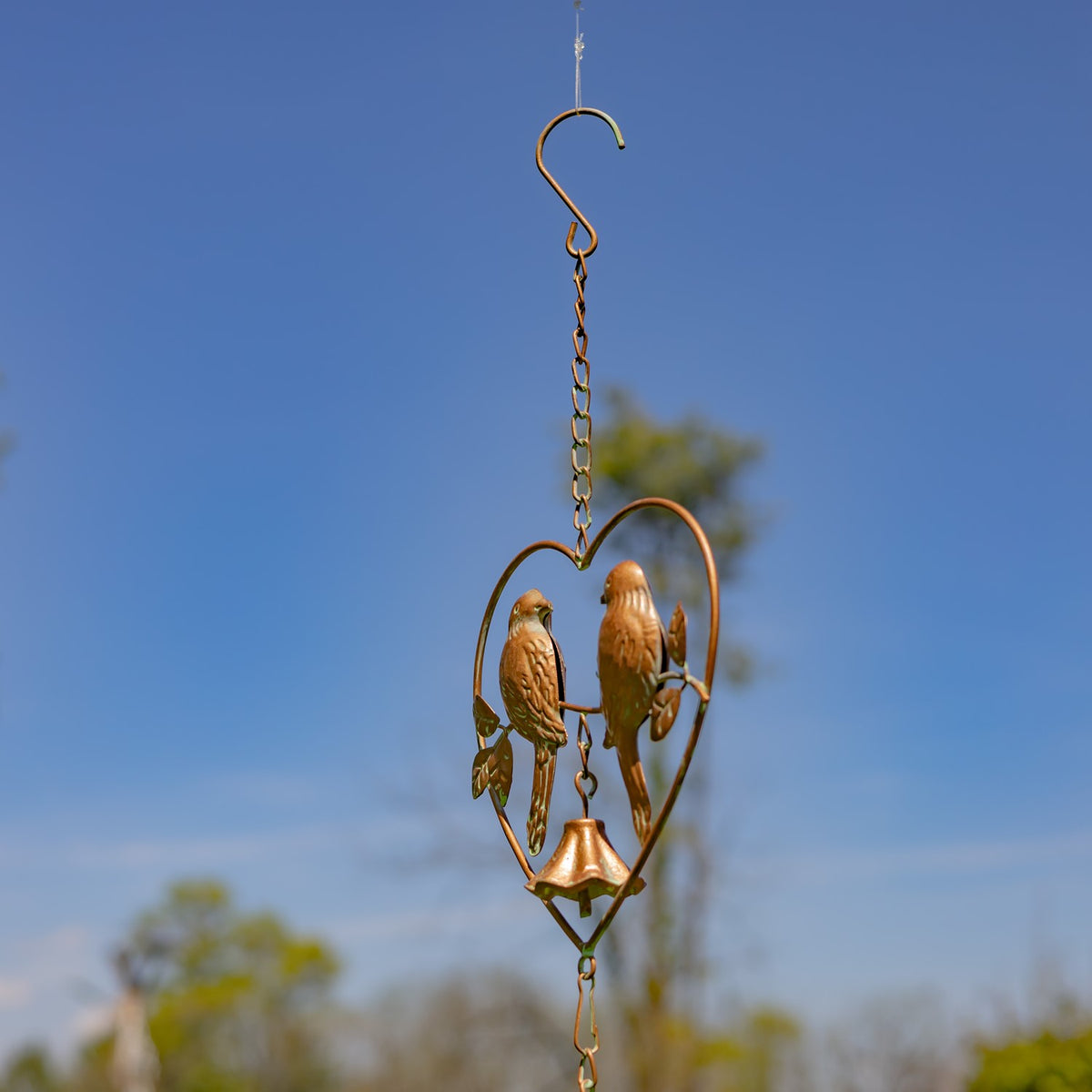 A heart-shaped bird ornament and bell hang from a chain and hook against a blue sky.