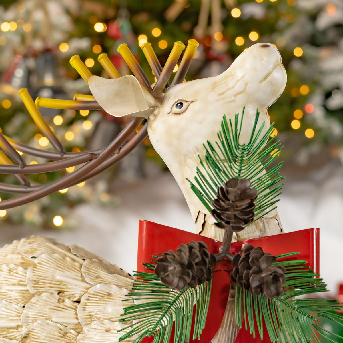 Close-up of an ivory reindeer with brown antlers, a red bow, and pinecones with green sprigs.