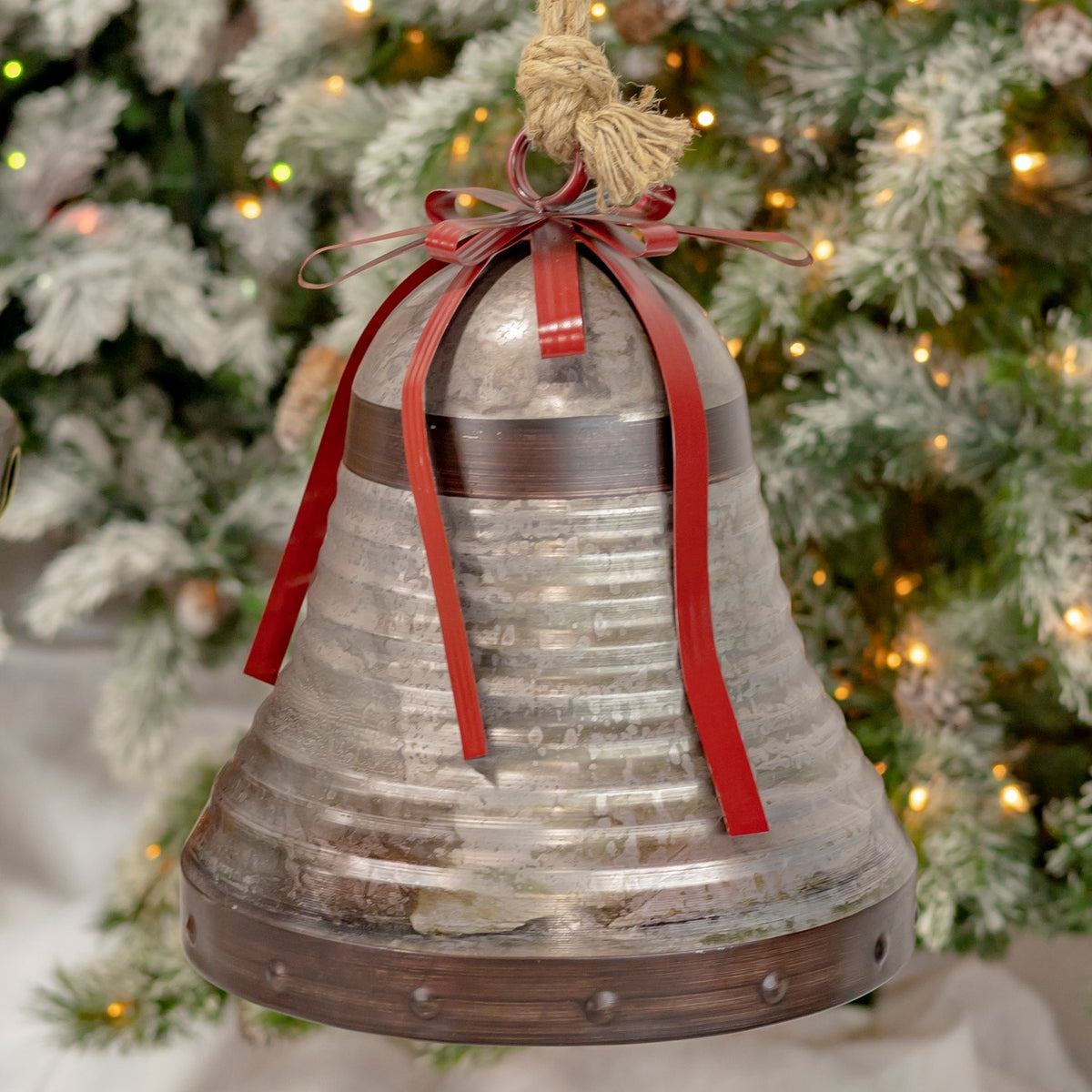 Tall, tiered galvanized bell with red ribbon streamers and a rope hanger in front of a tree.