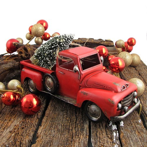 Angled view of a red pickup carrying a snowy miniature tree, surrounded by red and gold ornaments on wood.
