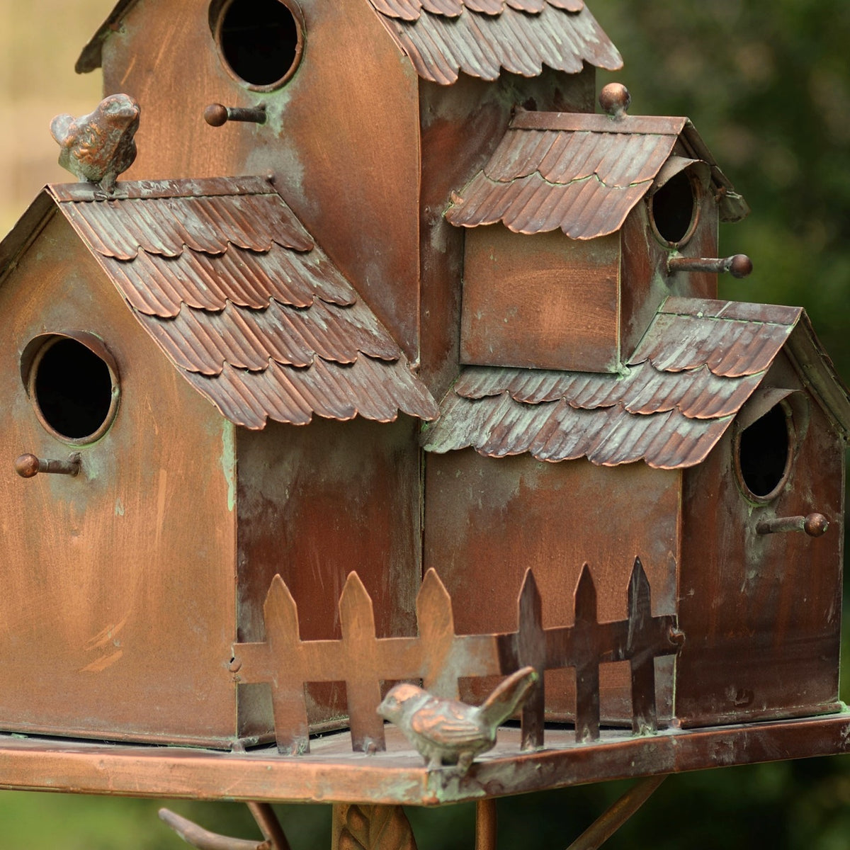 Birdhouse cluster close-up showing a miniature picket fence, bird figure, and weathered metal finish.