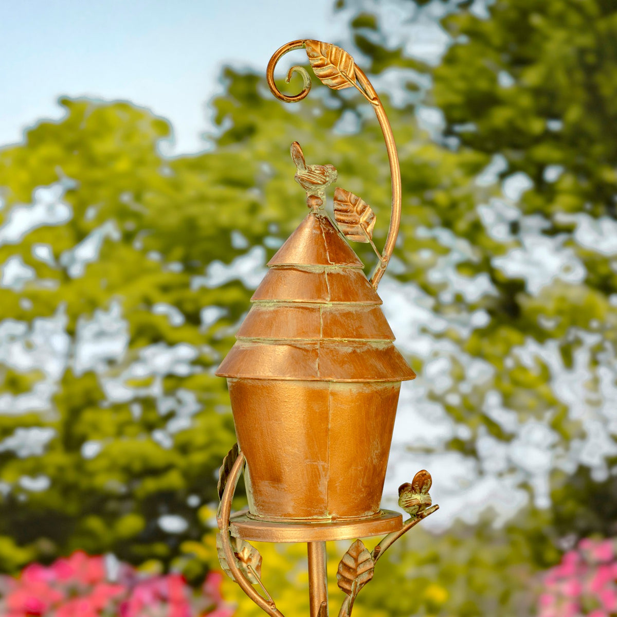 Back of the copper-toned birdhouse, showing its tiered roof, decorative birds, and curled leafy stake.