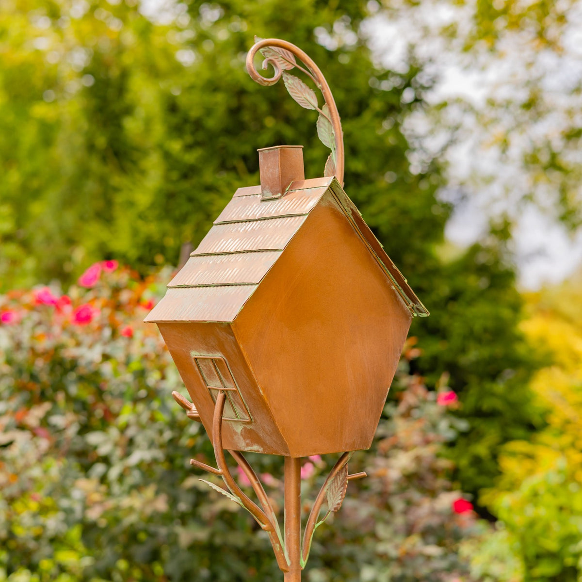 Antique copper-colored birdhouse garden stake with a peaked roof and curled metal accents among flowers.