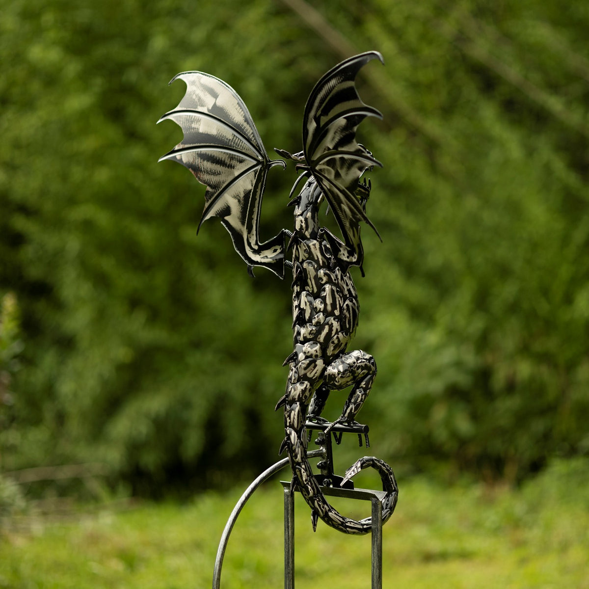 Metal dragon with raised wings perched atop its tall garden stake against greenery.