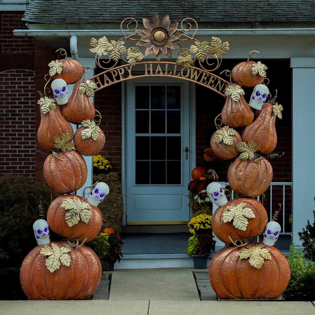 Pumpkin-and-skull Halloween arch stands across a front walkway in front of a house entrance.