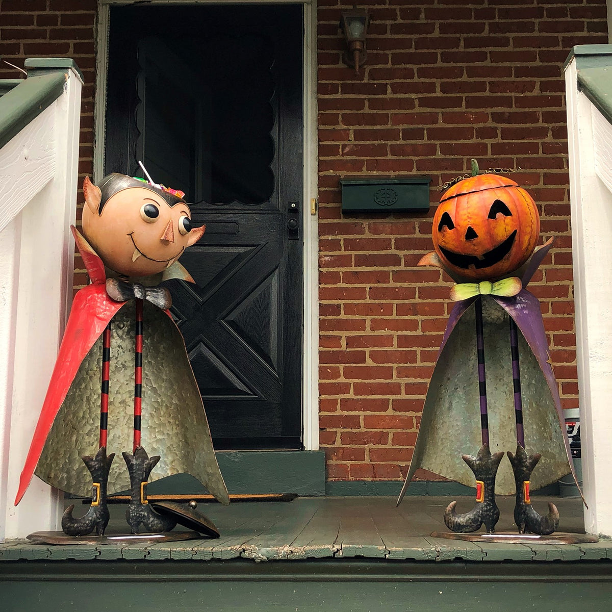 Pumpkin-headed figurine stands beside a red-caped character on a brick porch.