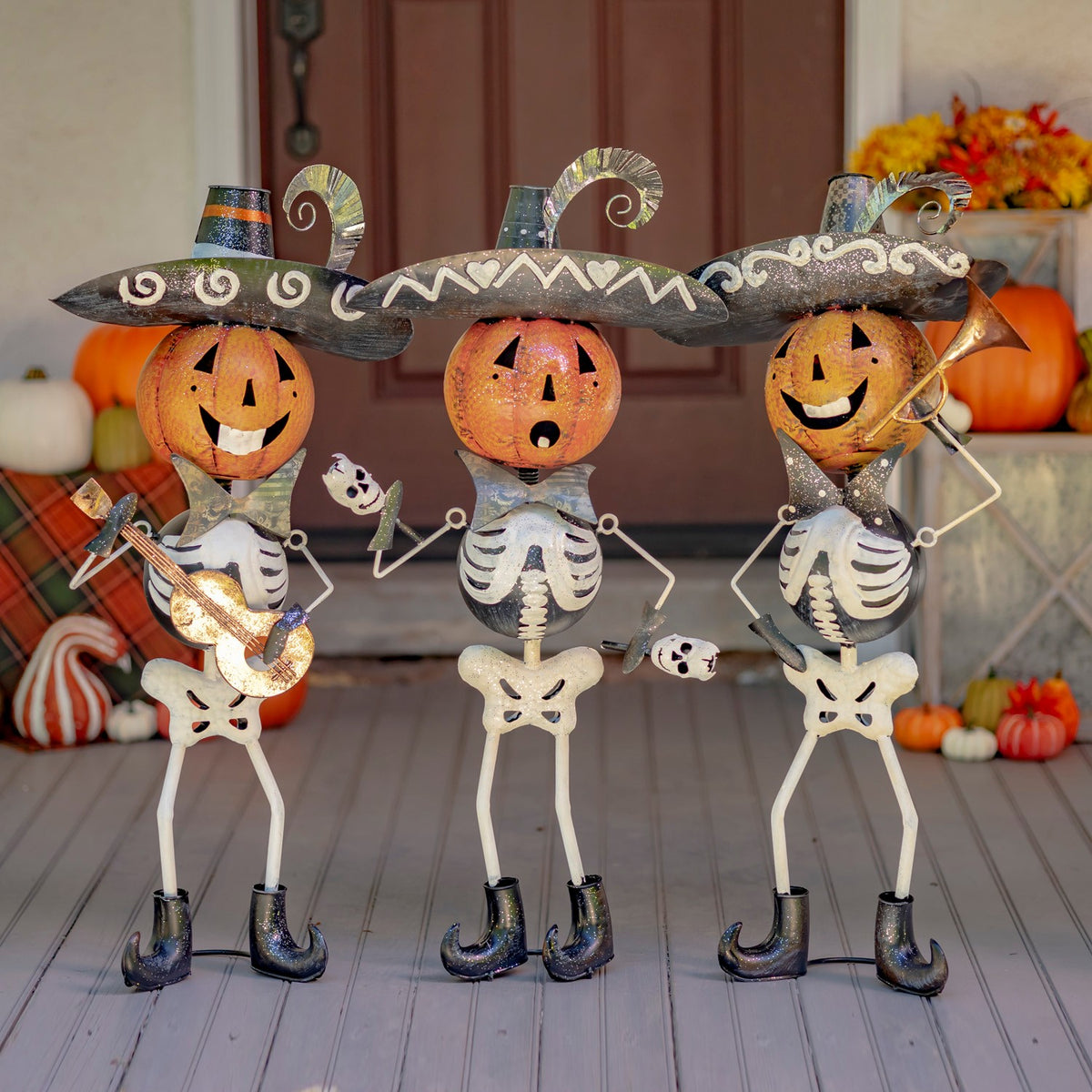 Three pumpkin-headed skeleton figurines in sombreros pose with instruments on a fall-decorated porch.