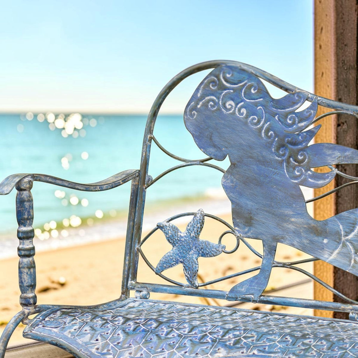 Close view of the bench’s mermaid backrest, starfish scrollwork, and patterned seat against a beach backdrop.
