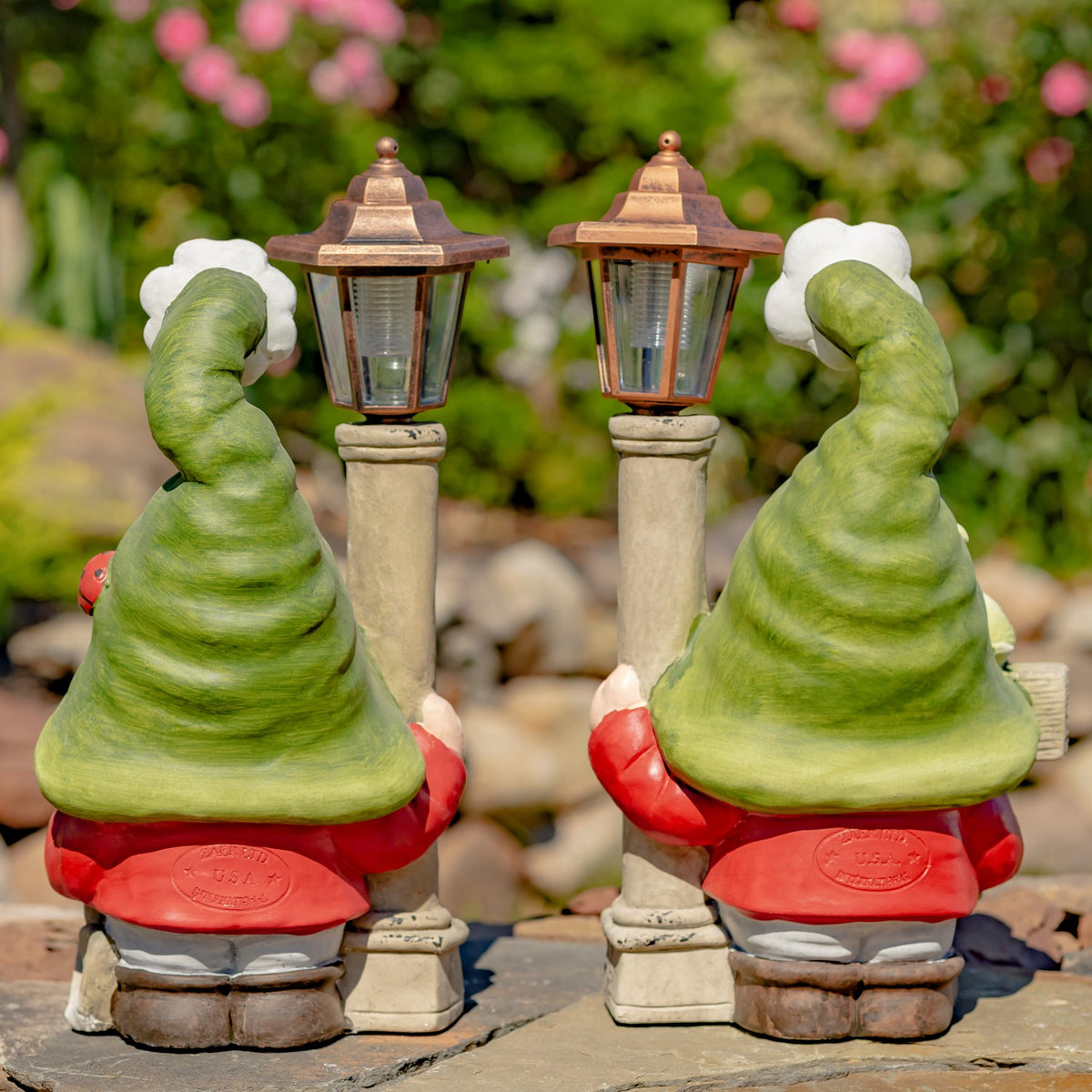 Two garden gnomes with tall lanterns, shown from behind among flowers and greenery.