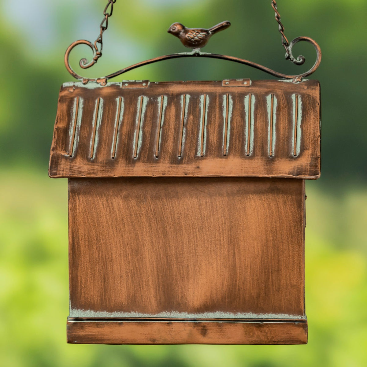 Back of the feeder with a plain copper-toned body, ridged roof, and decorative bird atop the hanging frame.