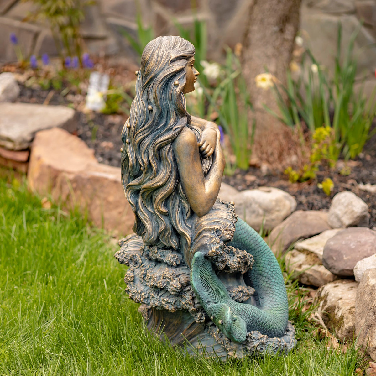 Side profile of the mermaid statue seated on a rock, with her tail curving down toward the grass.