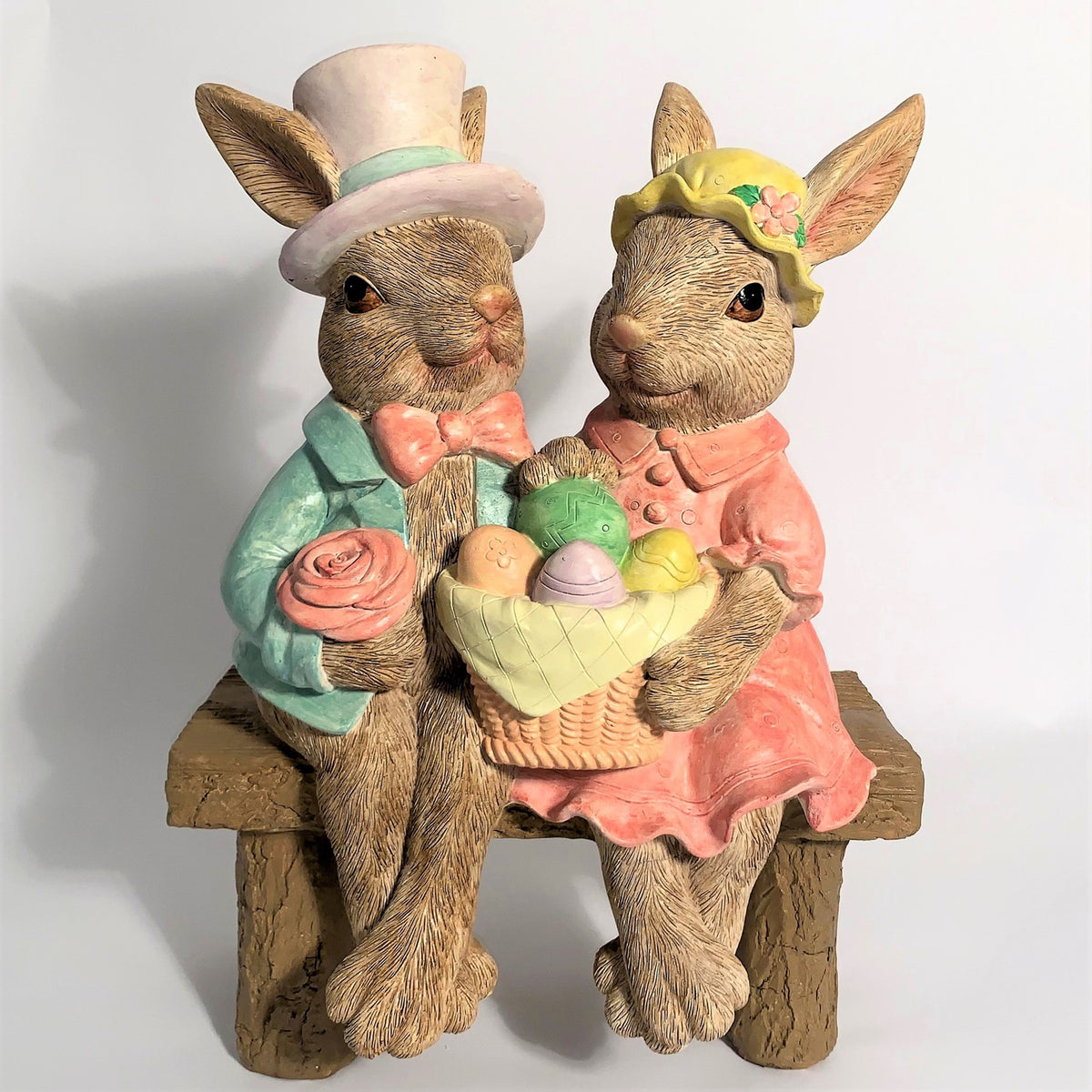 The rabbit couple faces forward on a rustic bench, with a basket of pastel eggs between them.