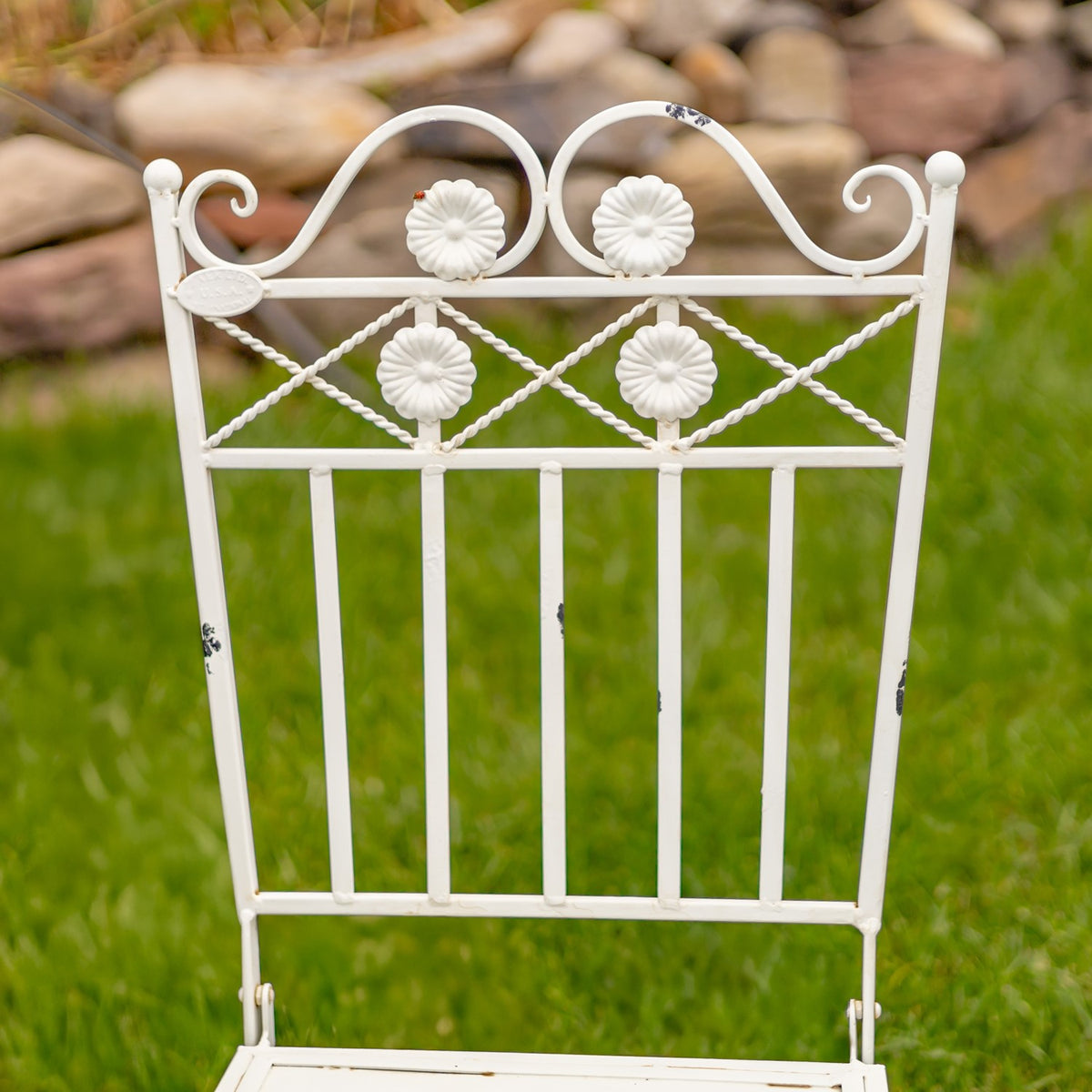 Pale metal chair back with curled scrollwork, flower ornaments, and vertical bars.
