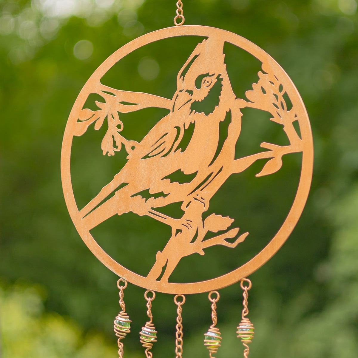Close-up of a copper-colored circular wind chime with a bird cutout and hanging metal strands against greenery.