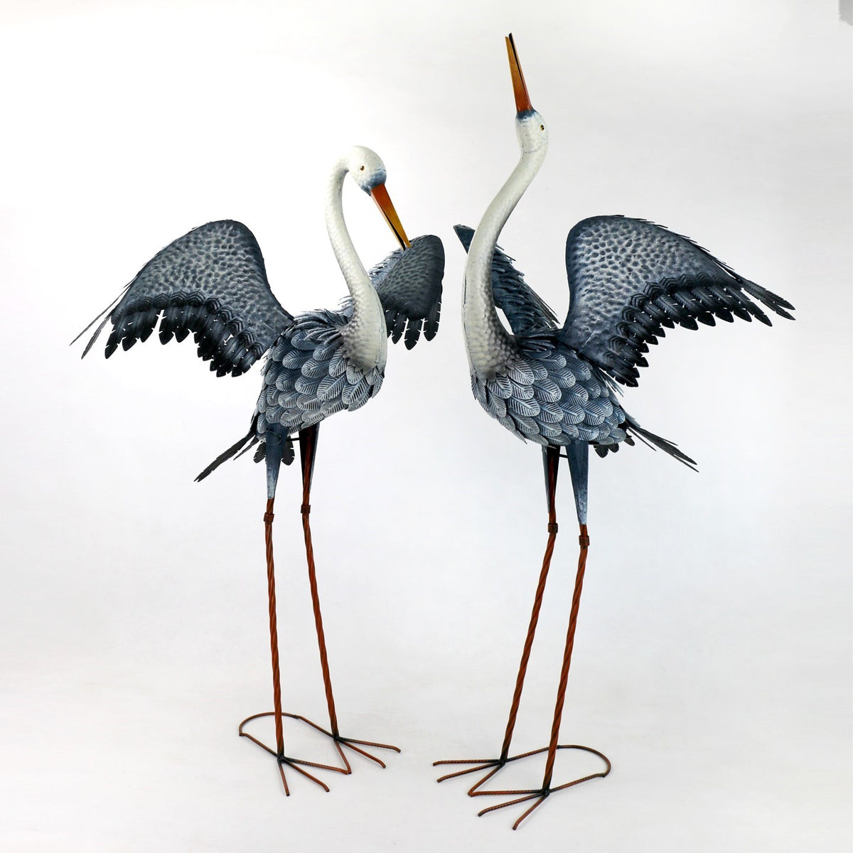 Two gray heron garden figurines stand side by side with wings spread and long metal legs.