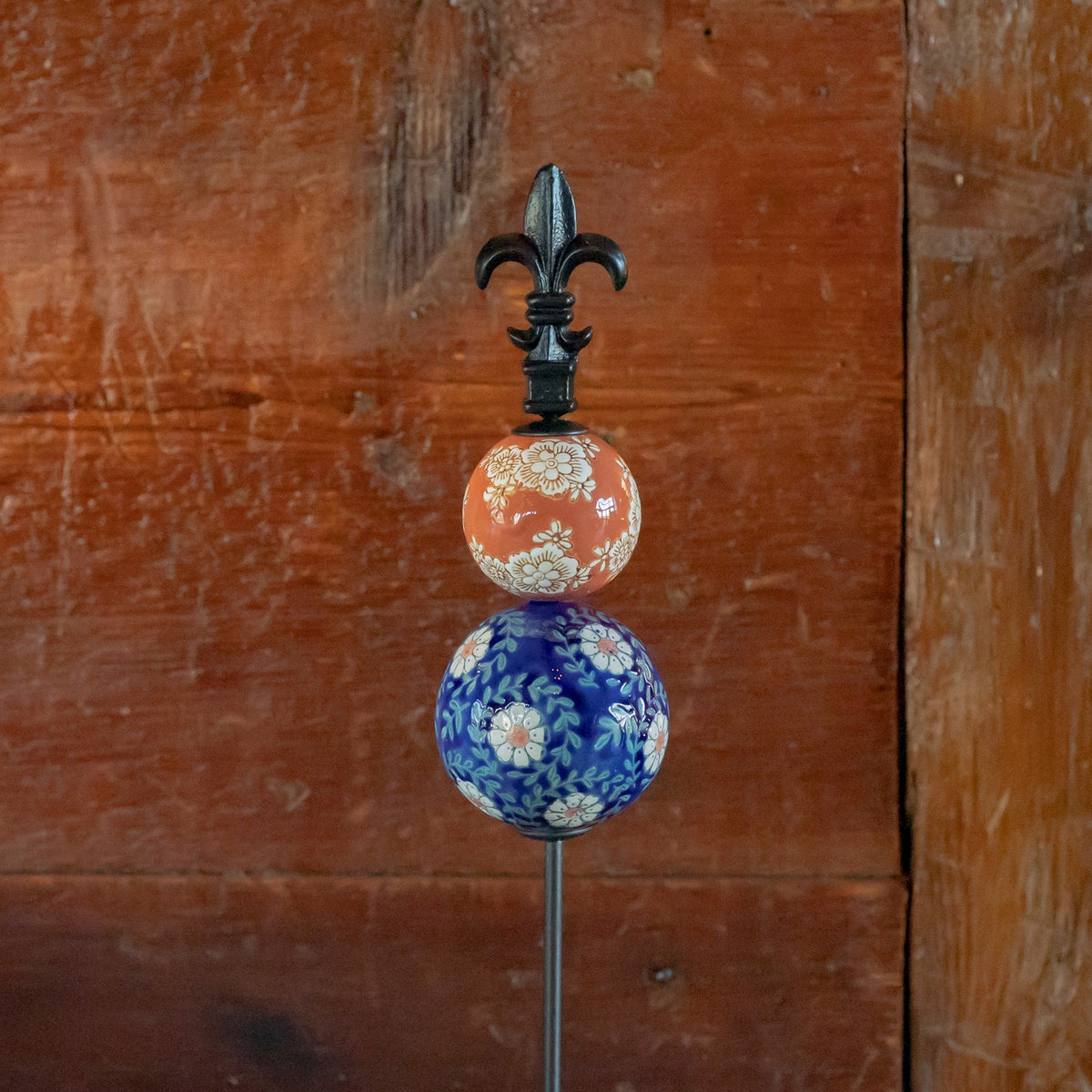 Close-up of a garden stake with two patterned ceramic balls and a black fleur-de-lis finial.