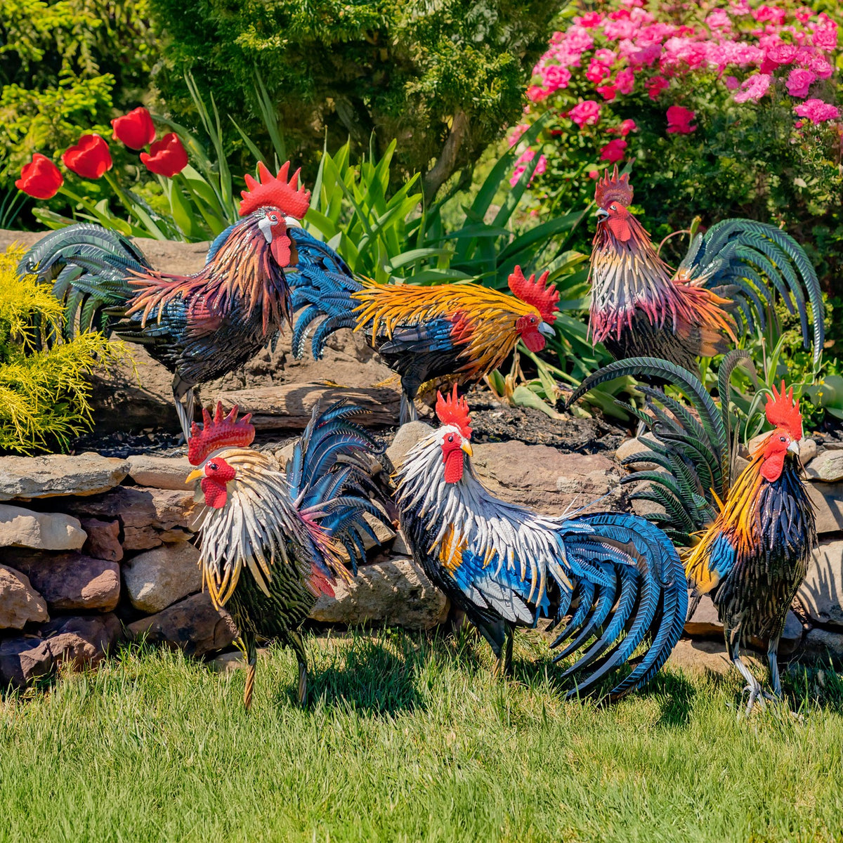 Several colorful metal rooster decorations arranged among flowers, rocks, and greenery.