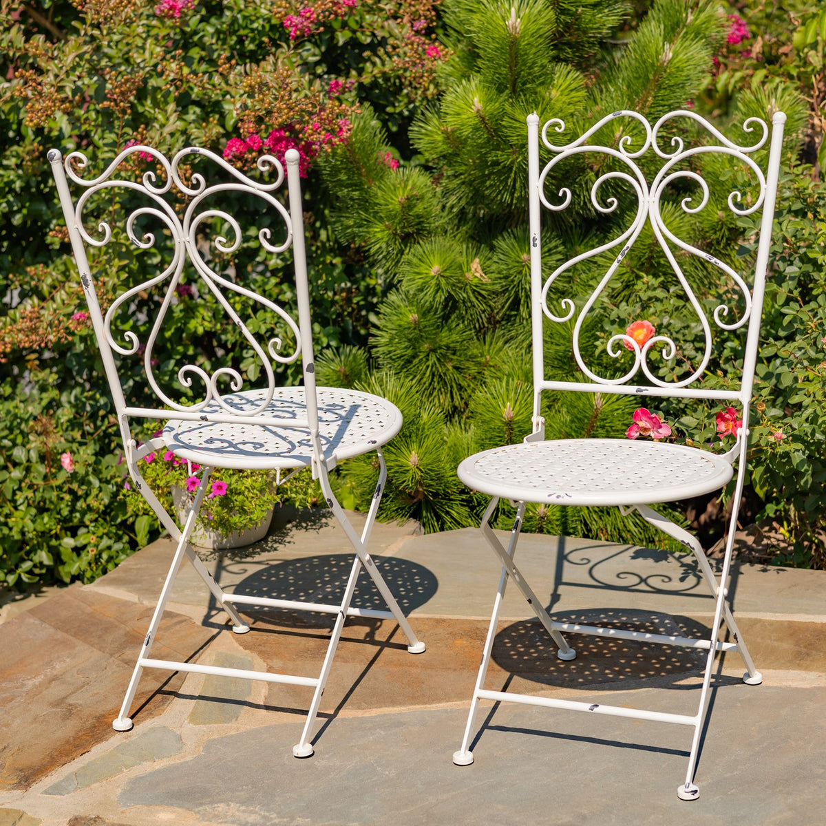 Two pale metal dining chairs with patterned seats and ornate scrollwork backs on a garden patio.