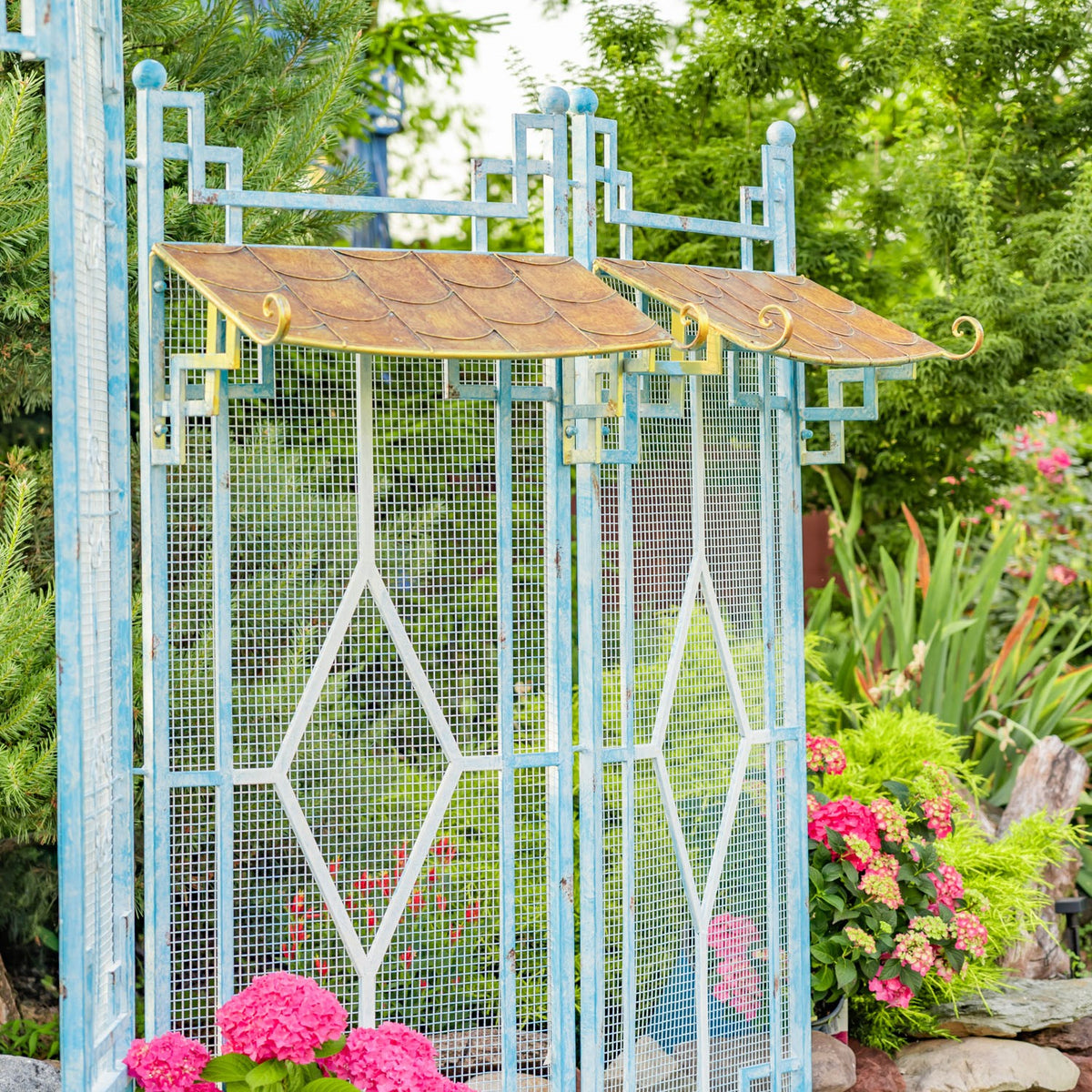 Blue garden gate with two mesh panels, diamond-patterned bars, pagoda roofs, and bright flowers nearby.
