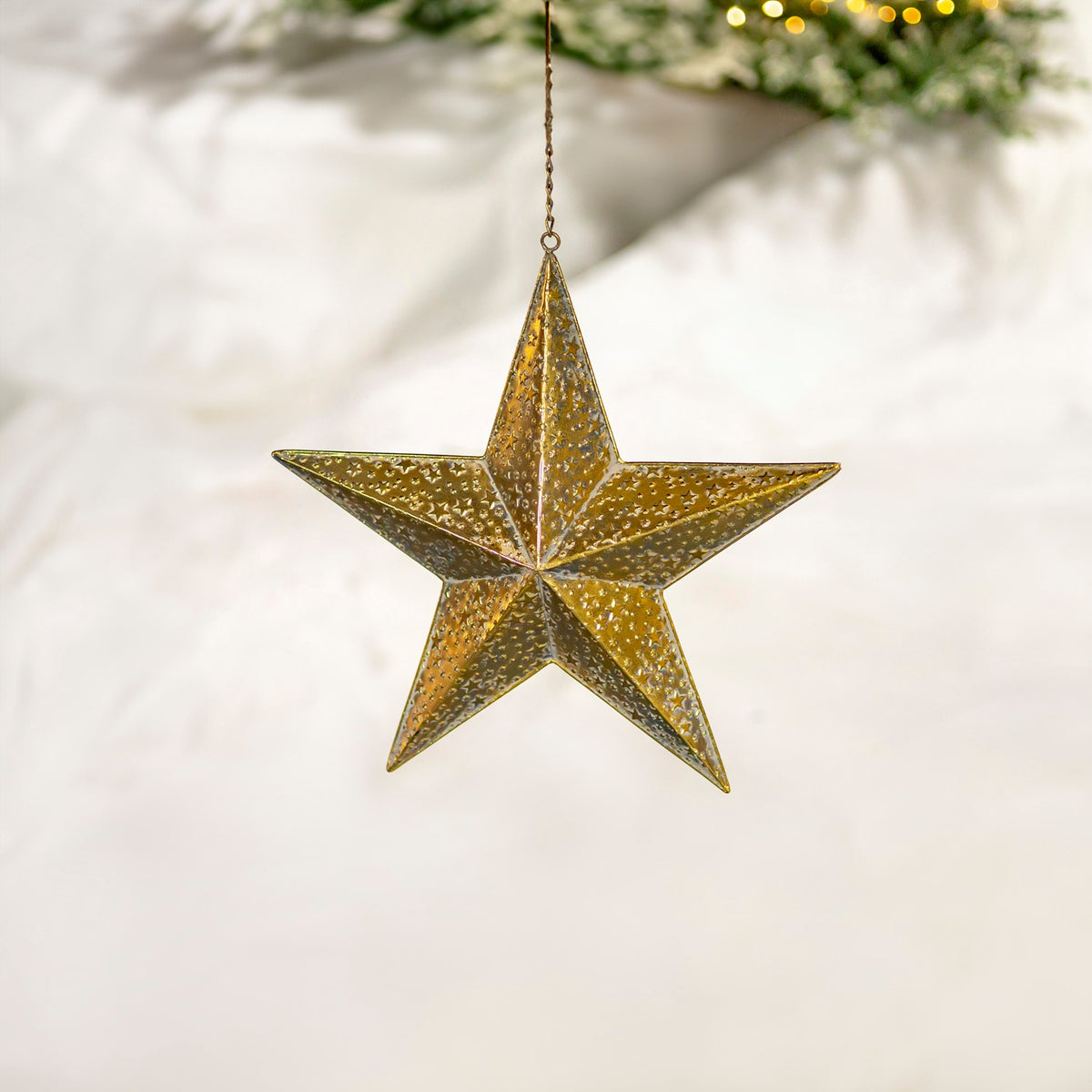 A hanging gold five-point star is shown against a pale backdrop with greenery and warm lights.