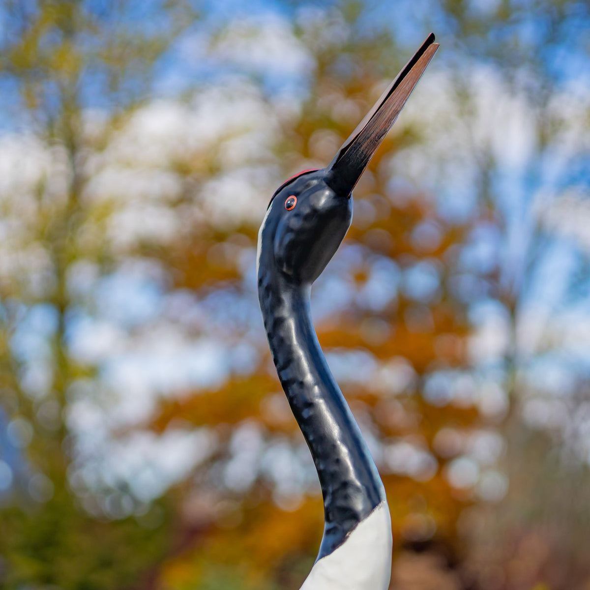 Close-up of a crane figurine’s curved dark neck, red eye, and long beak against blurred foliage.
