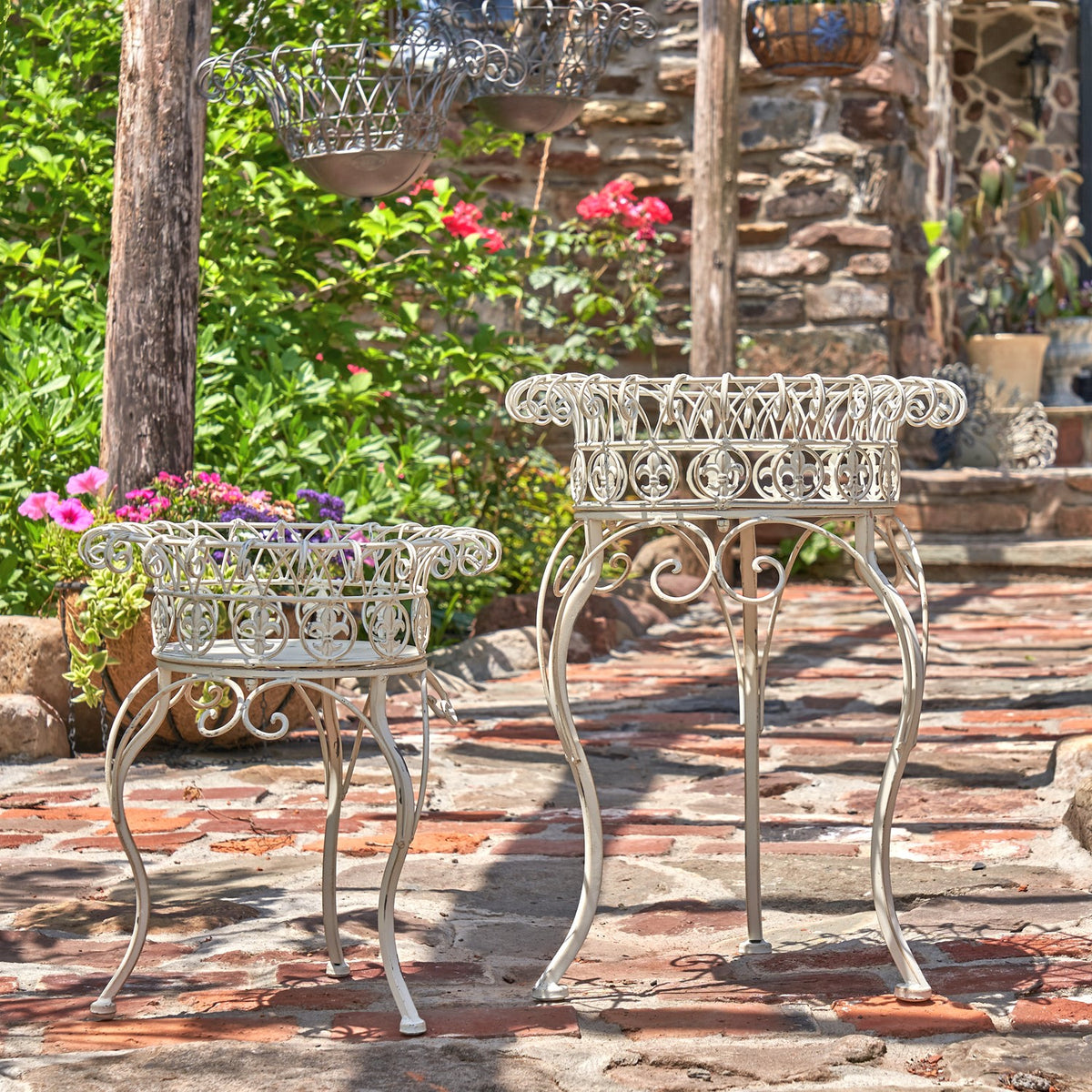 Two decorative metal plant stands on a patio, with flowers and hanging planters nearby.