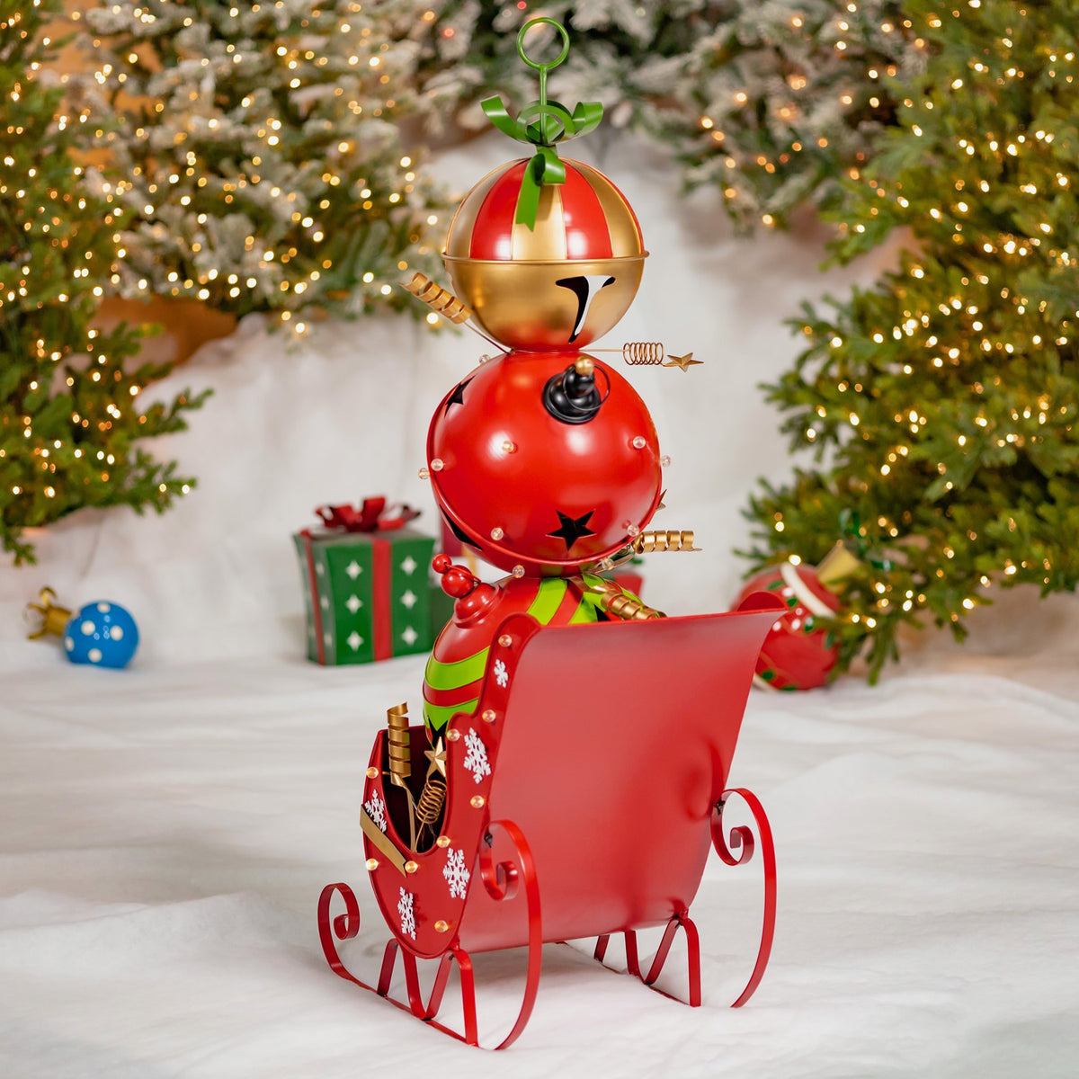 A red metal Christmas sleigh with curled runners and a festive figure sits beneath a lit tree.