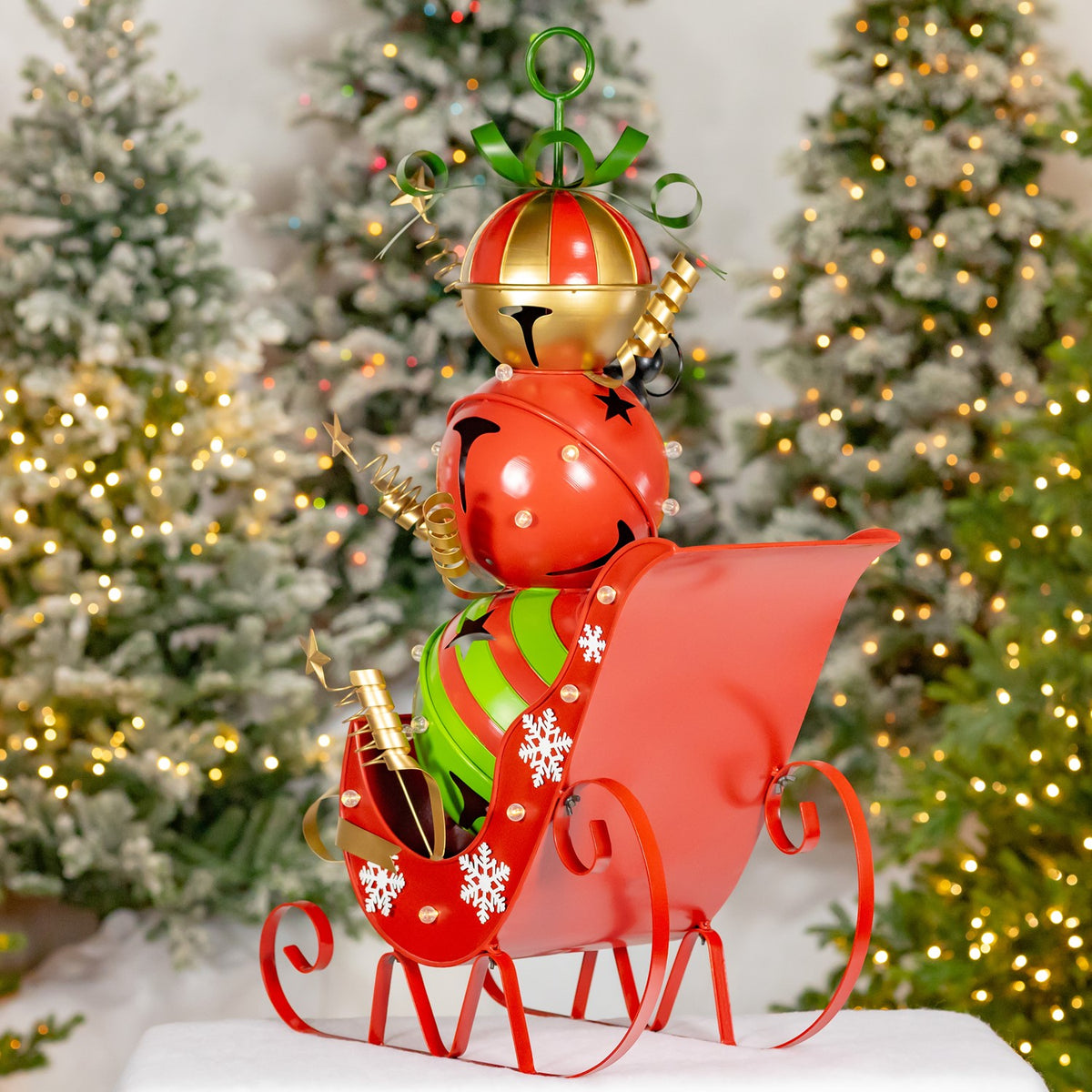 Red sleigh with curled runners and a tall curved back, carrying a festive bell figure beside lit trees.