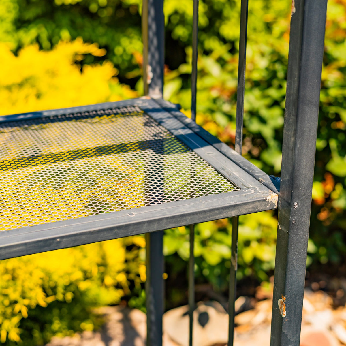 Close-up of a dark metal frame surrounding a perforated mesh shelf, with trees in the background.