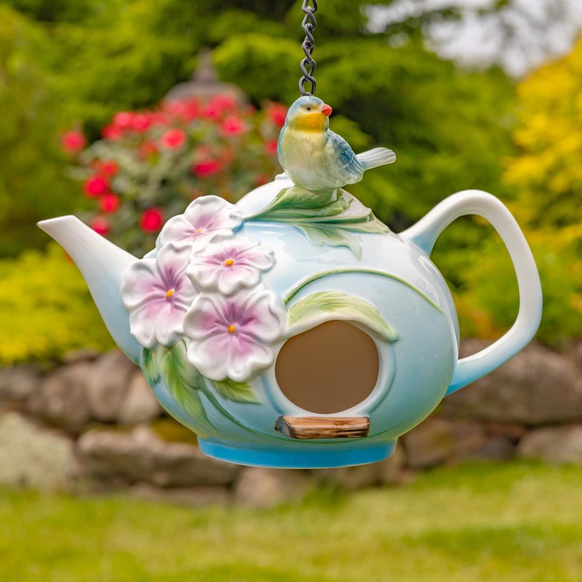 White and pale blue teapot birdhouse with raised pink blossoms and a perched bird, shown hanging in a garden.
