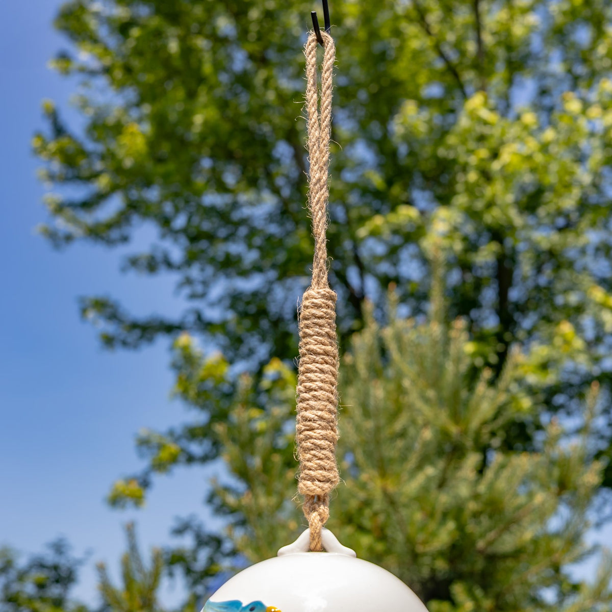 Close view of the birdhouse’s rope hanger and upper white portion against trees and blue sky.