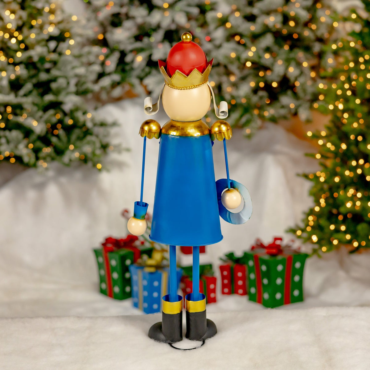 Blue-coated nutcracker figurine wearing a red hat and gold crown, with dangling ornaments and a festive tree backdrop.