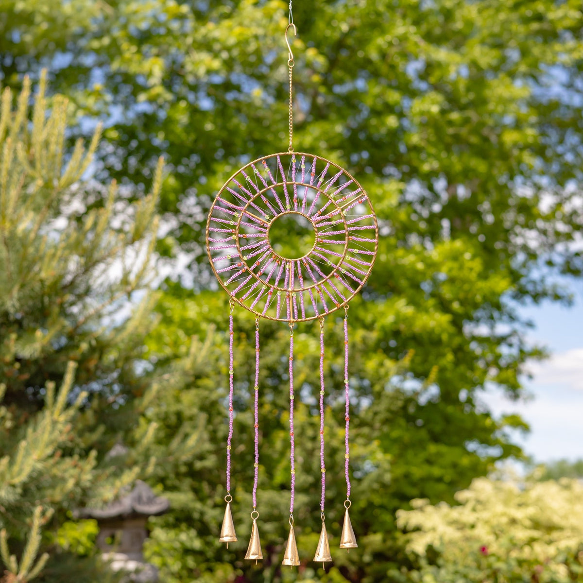 Pink and purple beaded wind chime with a circular top and dangling bells, hanging among leafy trees.