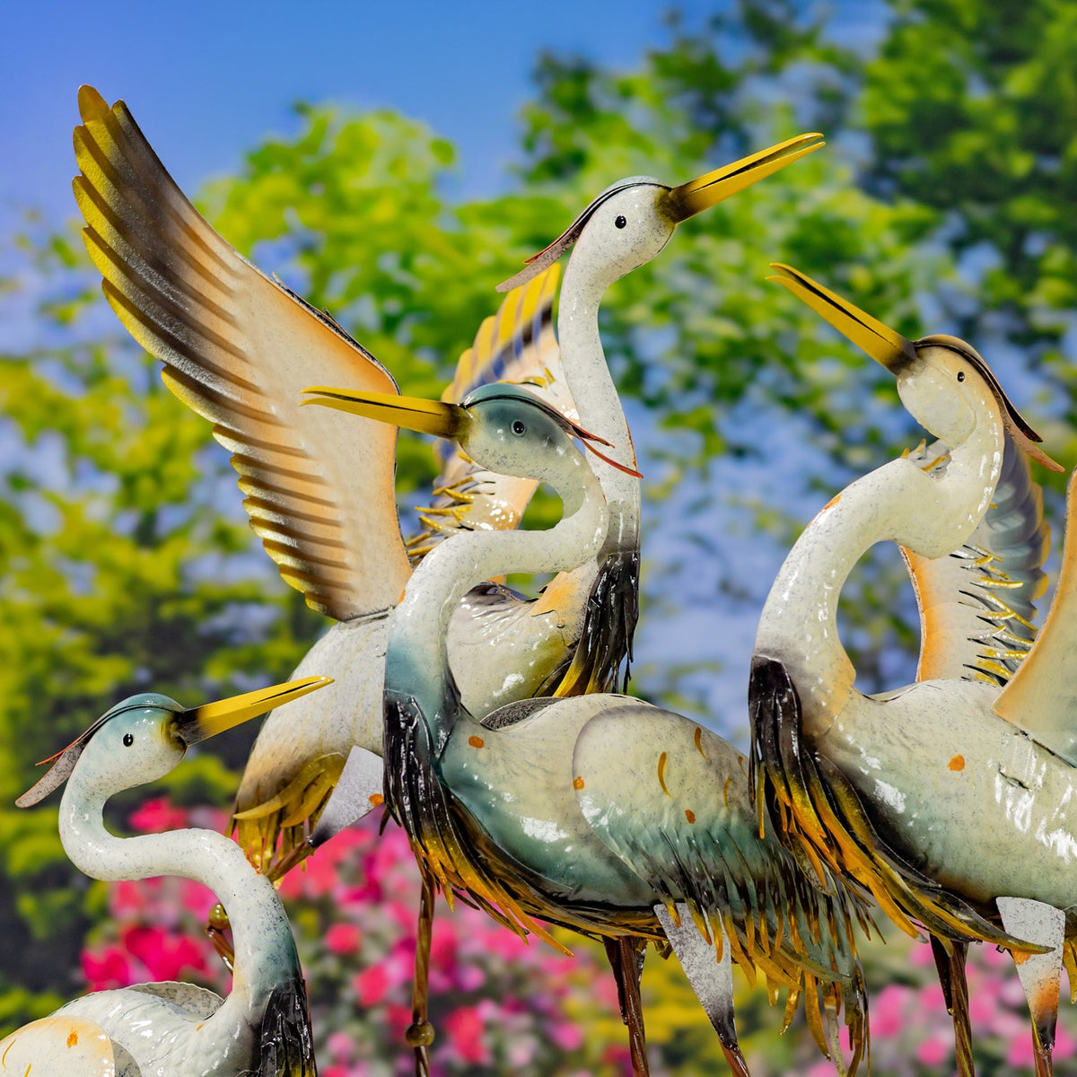 Several metal crane garden figurines grouped together, including one with raised wings, against trees and flowers.