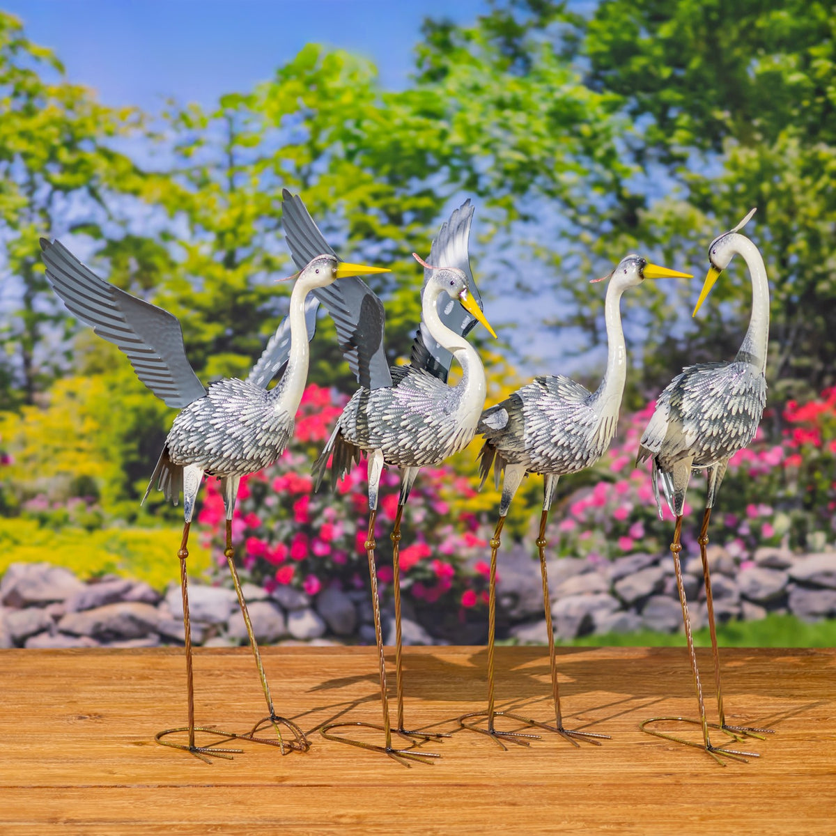 Four metal heron figurines, including two with raised wings, are arranged in a row against a garden backdrop.