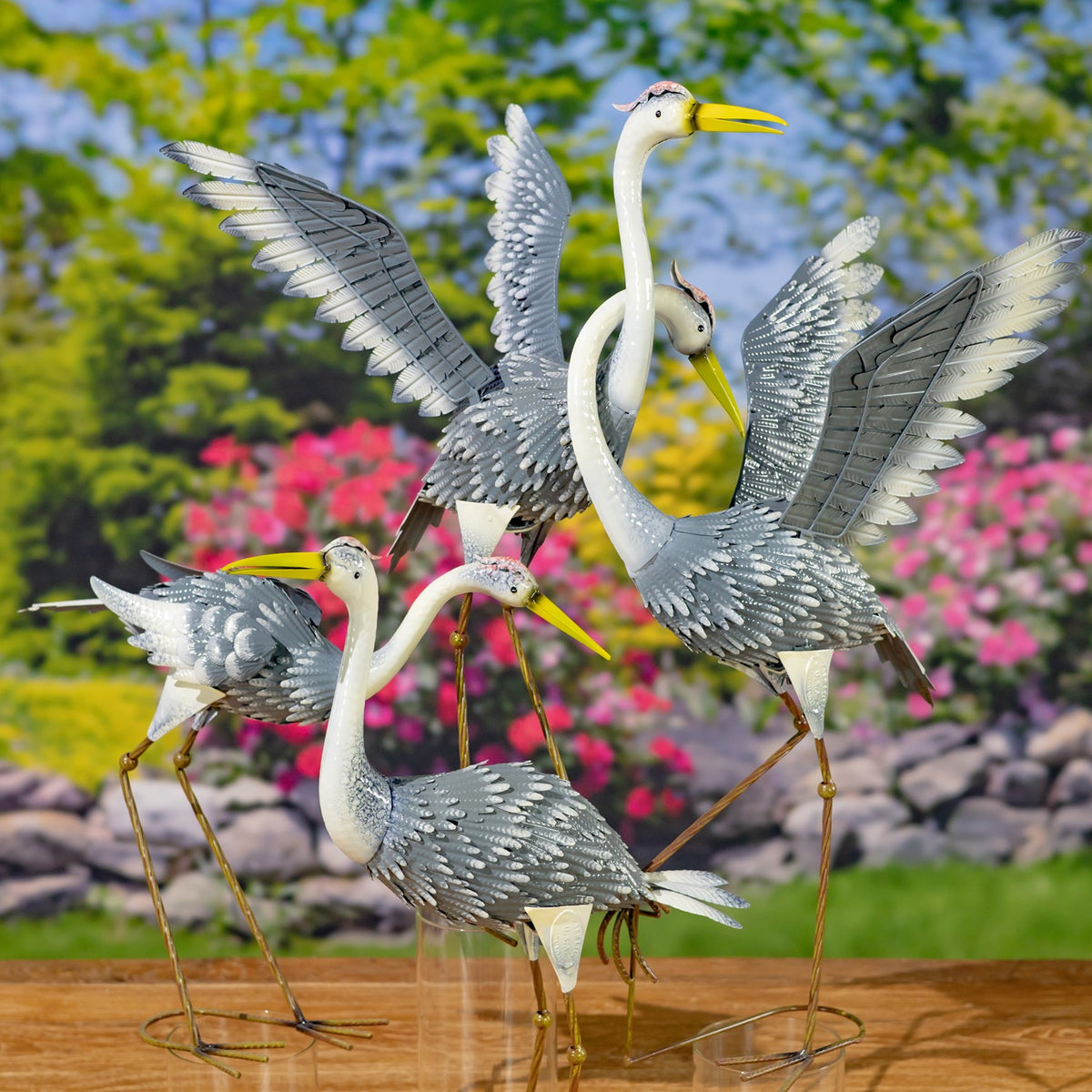 Four metal heron figurines in varied poses, including birds with raised wings, arranged together in a garden setting.
