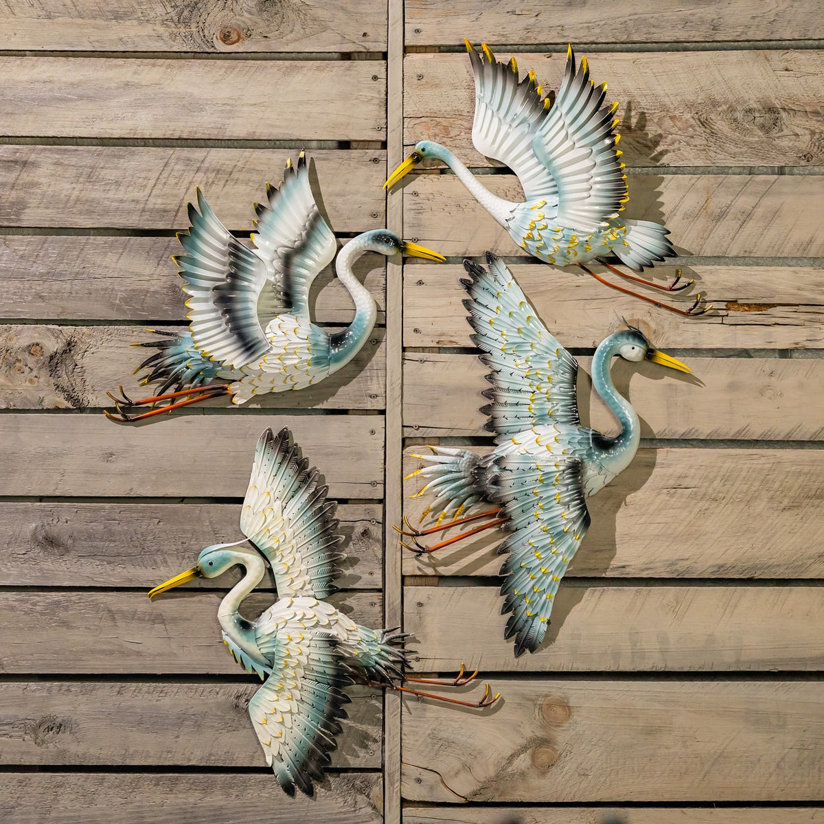 Four metal crane decorations in varied flying poses displayed together on a rustic plank wall.