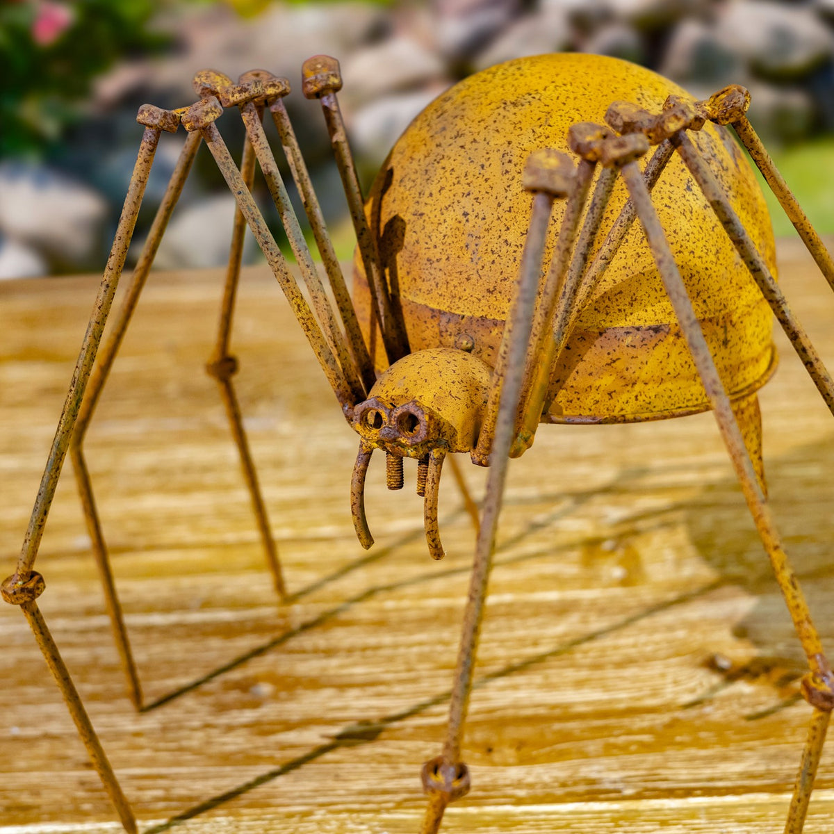 Rust-finished metal spider figurine on a wooden surface, shown close up against a garden backdrop.