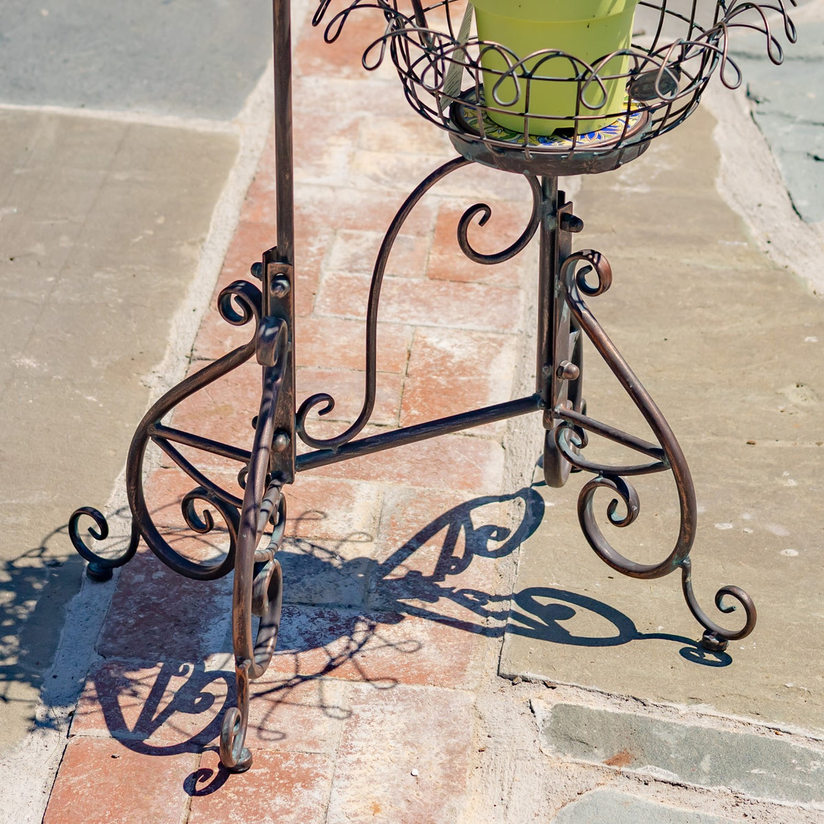 Curled metal legs and crossbars beneath a wire basket holding a green plant pot on a stone patio.
