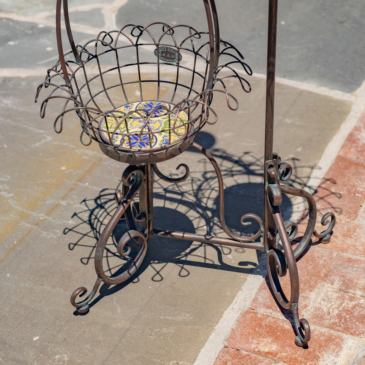 The wire basket sits above a bronze-colored stand with curved scrollwork legs on stone paving.