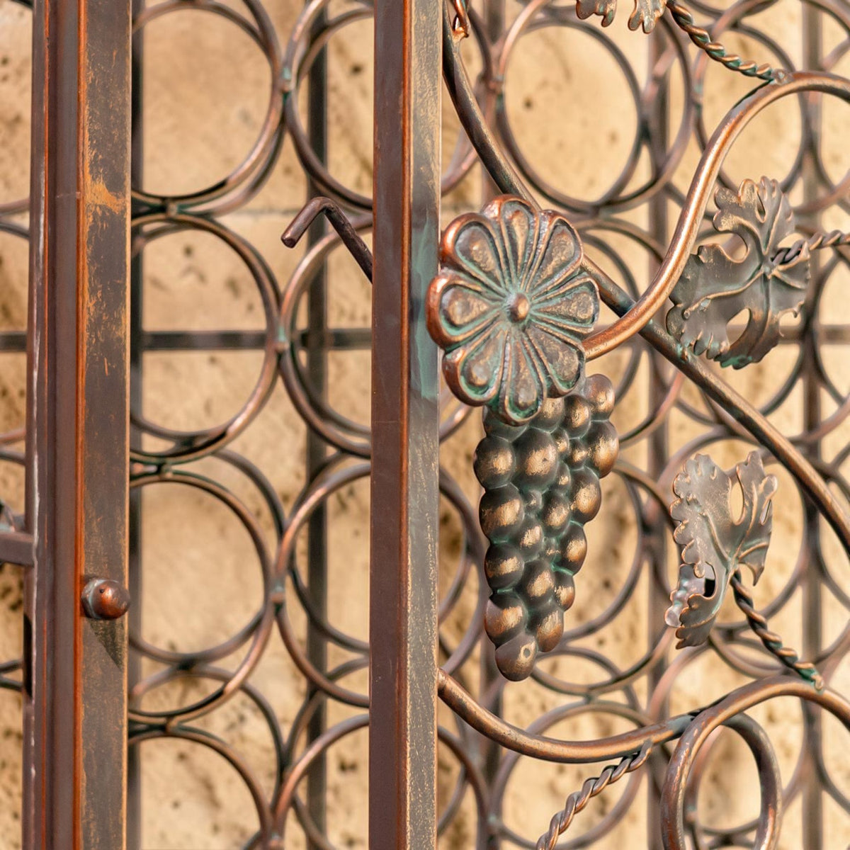Side view of the iron rack’s circular bottle holders and grapevine ornament against a stone wall.