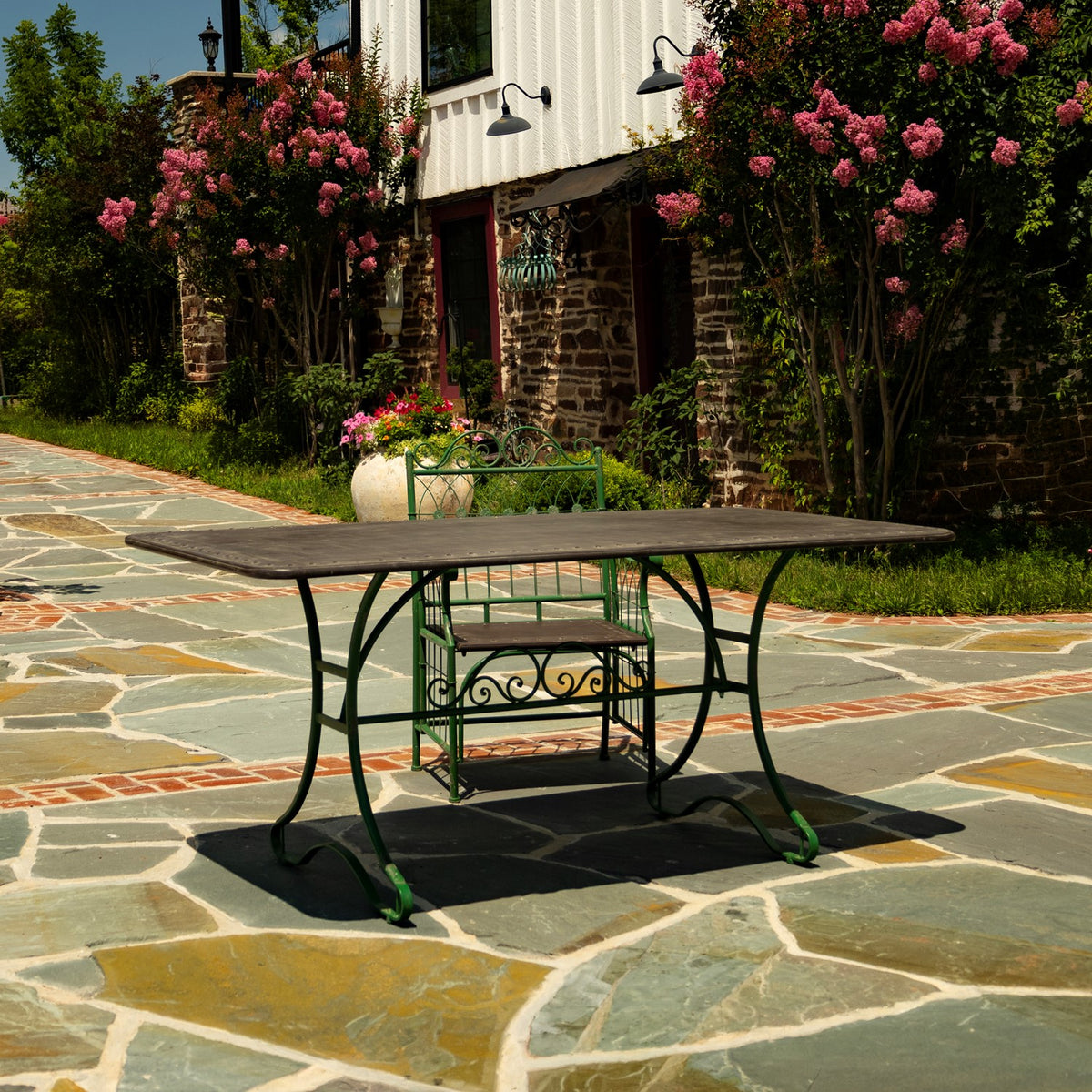 Green metal dining table and matching chair on a stone patio beside a flower-lined building