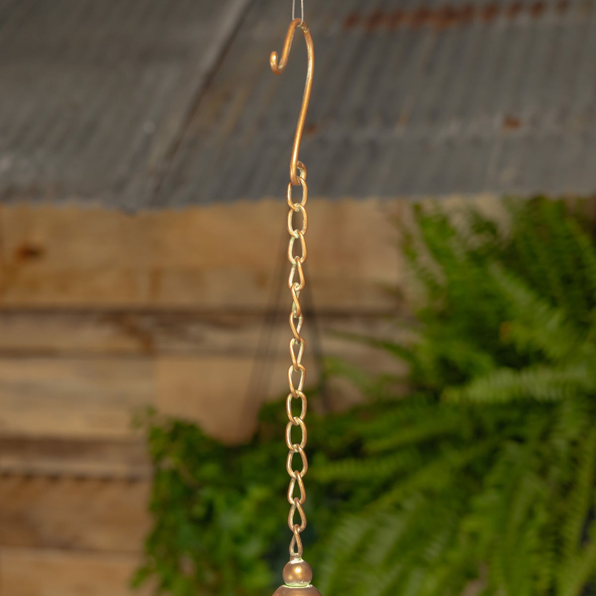 A gold-toned chain and hook hang against a wooden fence, with the top of the birdhouse just visible below.