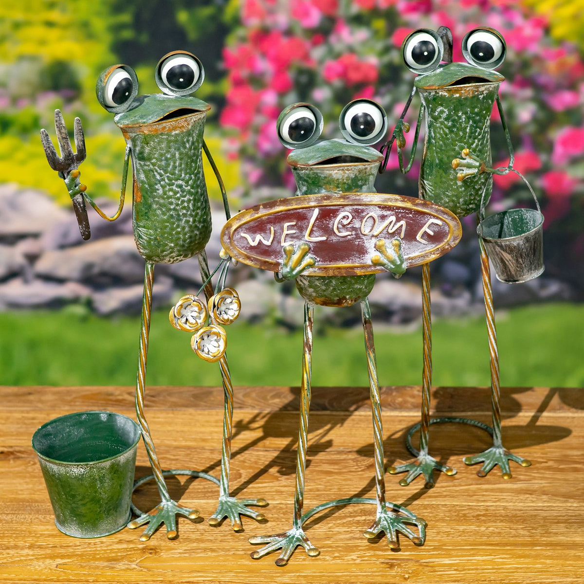 Green metal frog figures surround an oval garden sign, with a small pot in the foreground.