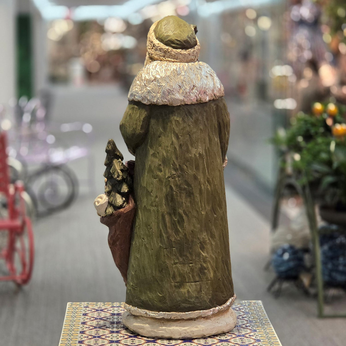Back of a carved Santa figurine in a long green coat, with a small tree and sack visible at his side.